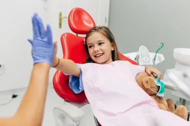 Uśmiechnięta dziewczynka w fotelu dentystycznym przybija piątkę w rękawiczce, trzyma misia.