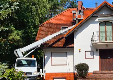 Mężczyzna na podnośniku myje dach domu z czerwoną dachówką.