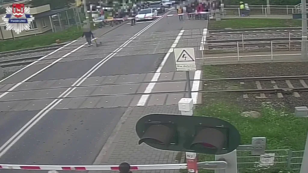 Przejazd kolejowy w Piasecznie z opuszczonymi szlabanami, piesi i pojazdy czekają za torami.