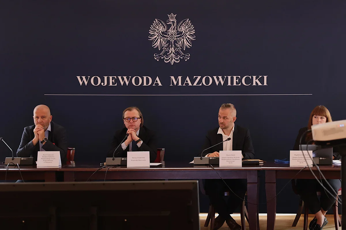 Cztery osoby siedzą za stołem konferencyjnym w sali z napisem „Wojewoda Mazowiecki” i orłem na ścianie.