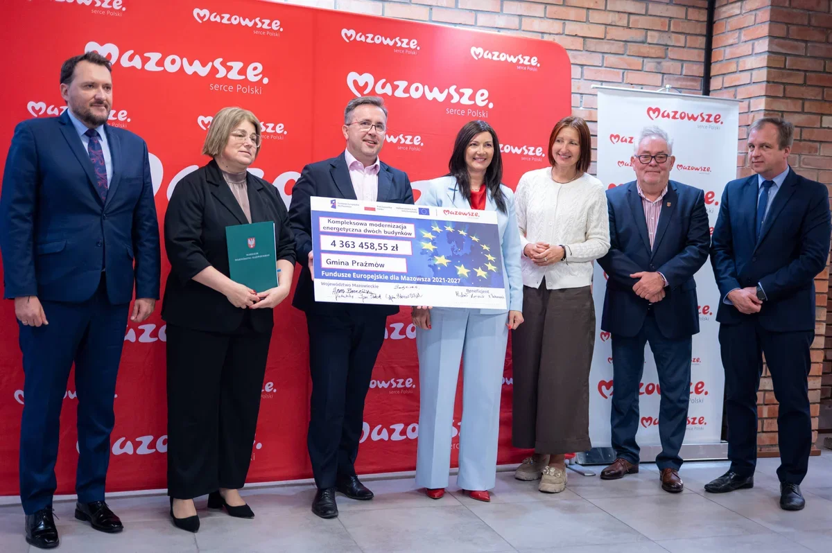 Grupa osób podczas uroczystego wręczenia symbolicznego czeku na modernizację energetyczną budynków dla Gminy Prażmów, na tle czerwonej ścianki z logo Mazowsze.