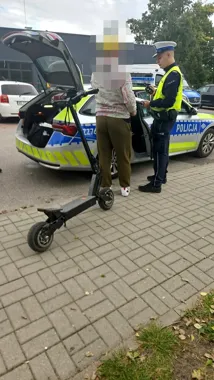 Policjant w kamizelce odblaskowej wypisuje mandat kobecie stojącej przy policyjnym radiowozie, obok na chodniku stoi elektryczna hulajnoga.