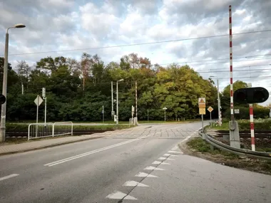 Przejazd kolejowy przez drogę asfaltową w otoczeniu zieleni, z szlabanami i sygnalizacją świetlną, w słoneczny dzień z zachmurzonym niebem.