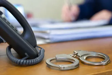 Kajdanki i telefon leżą na drewnianym biurku, w tle rozmyte ręce osoby przy stosie dokumentów.