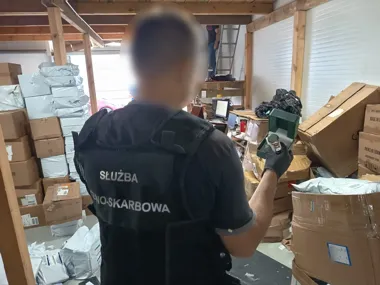 Funkcjonariusz Służby Celno-Skarbowej w pomieszczeniu magazynowym wypełnionym paczkami i kartonami ogląda przedmiot w zielonym pudełku.