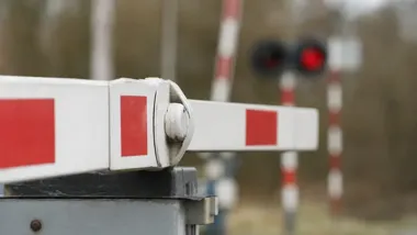Opuszczone zapory na przejeździe kolejowym z czerwonym sygnałem świetlnym w tle.