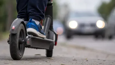 Zbliżenie na tylne koło hulajnogi elektrycznej i stopa dziecka w niebieskim bucie, w tle rozmyte auto z włączonymi światłami na drodze.